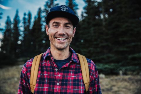 Man outside on a hike wearing a red flannel shirt, yellow backpack and ballcap smiling at the camera